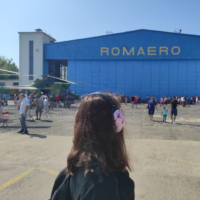 imagine cu o fata care poarta casti antifonice iar in spatele ei este hangarul Romaero Baneasa