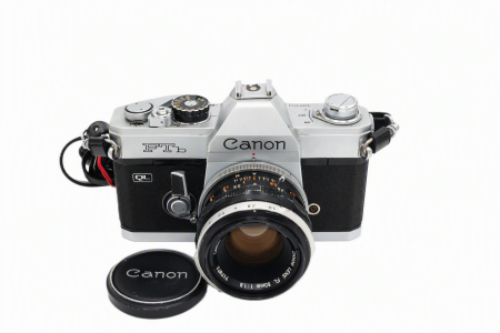 Aparate foto SLR (pe film) (SH) - Kit aparat foto SLR pe film 35mm Canon FTb QL second hand în finisaj argintiu, cu obiectivul Canon Lens FL 50mm F1.8 montat și capacul frontal așezat alături pe fundal alb izolat.