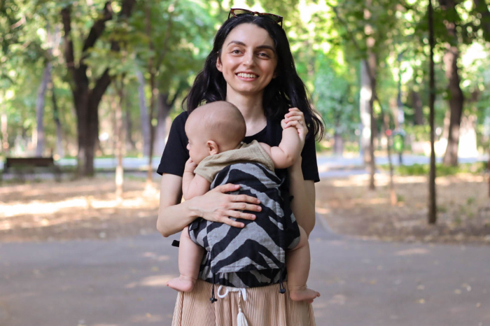 Marsupiu ergonomic Luna Una de culoare neagra, purtat de un părinte cu bebeluș. Design ajustabil, bretele moi și glugă pentru susținerea capului. [8]
