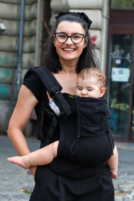 Marsupiu ergonomic Luna Una de culoare neagra, purtat de un părinte cu bebeluș. Design ajustabil, bretele moi și glugă pentru susținerea capului. [1]