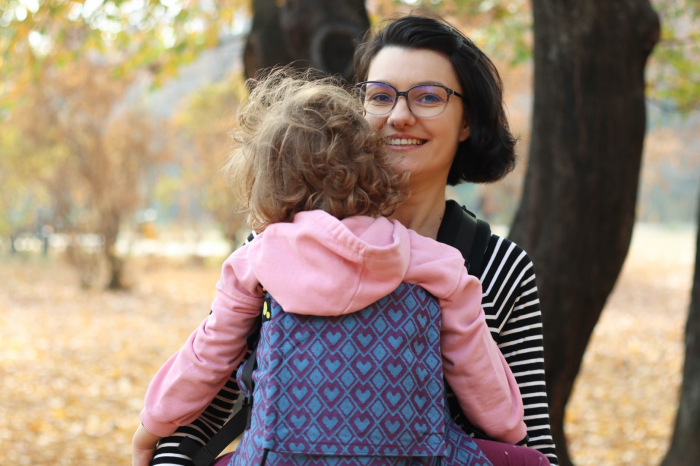Marsupiu ergonomic Luna Una de culoare neagra, purtat de un părinte cu bebeluș. Design ajustabil, bretele moi și glugă pentru susținerea capului. [4]