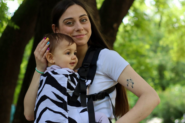 Marsupiu ergonomic Luna Una de culoare neagra, purtat de un părinte cu bebeluș. Design ajustabil, bretele moi și glugă pentru susținerea capului. [3]
