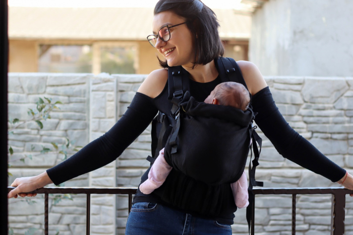 Marsupiu ergonomic Luna Una de culoare neagra, purtat de un părinte cu bebeluș. Design ajustabil, bretele moi și glugă pentru susținerea capului. [4]