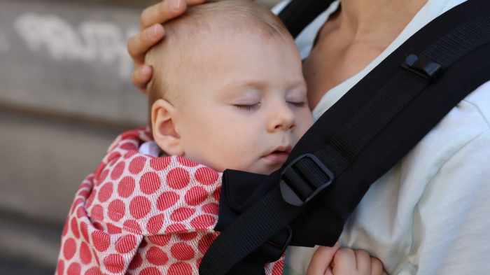 Marsupiu ergonomic Luna Una de culoare neagra, purtat de un părinte cu bebeluș. Design ajustabil, bretele moi și glugă pentru susținerea capului. [4]