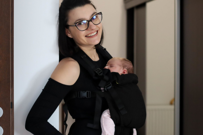 Marsupiu ergonomic Luna Una de culoare neagra, purtat de un părinte cu bebeluș. Design ajustabil, bretele moi și glugă pentru susținerea capului. [5]