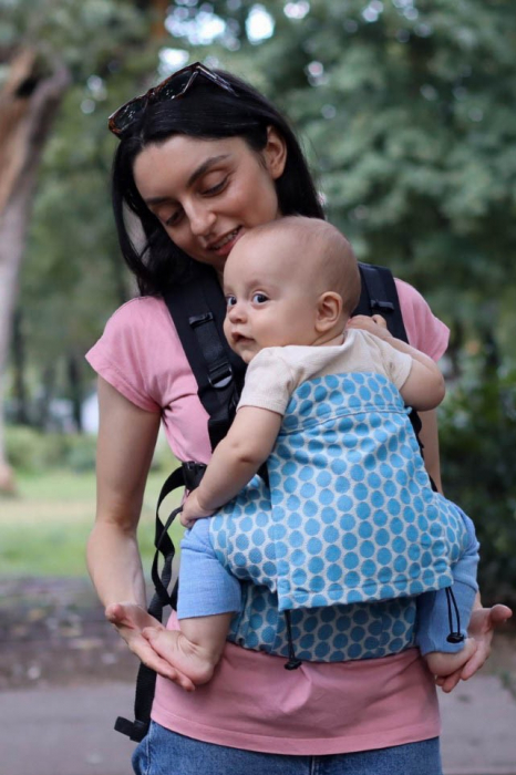 Marsupiu ergonomic Luna Una de culoare neagra, purtat de un părinte cu bebeluș. Design ajustabil, bretele moi și glugă pentru susținerea capului. [1]