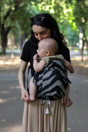 Marsupiu ergonomic Luna Una de culoare neagra, purtat de un părinte cu bebeluș. Design ajustabil, bretele moi și glugă pentru susținerea capului. [6]