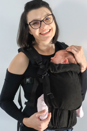 Marsupiu ergonomic Luna Una de culoare neagra, purtat de un părinte cu bebeluș. Design ajustabil, bretele moi și glugă pentru susținerea capului. [1]