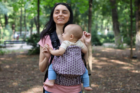 Marsupiu ergonomic Luna Una de culoare neagra, purtat de un părinte cu bebeluș. Design ajustabil, bretele moi și glugă pentru susținerea capului. [2]