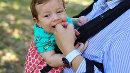 Marsupiu ergonomic Luna Una de culoare neagra, purtat de un părinte cu bebeluș. Design ajustabil, bretele moi și glugă pentru susținerea capului. [1]