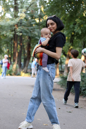 Marsupiu ergonomic Luna Una de culoare neagra, purtat de un părinte cu bebeluș. Design ajustabil, bretele moi și glugă pentru susținerea capului. [2]