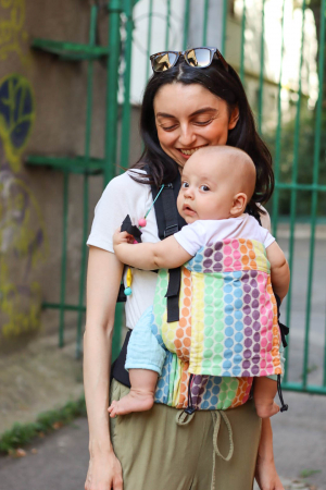 Marsupiu ergonomic Luna Una de culoare neagra, purtat de un părinte cu bebeluș. Design ajustabil, bretele moi și glugă pentru susținerea capului. [1]