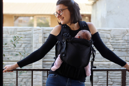 Marsupiu ergonomic Luna Una de culoare neagra, purtat de un părinte cu bebeluș. Design ajustabil, bretele moi și glugă pentru susținerea capului. [3]