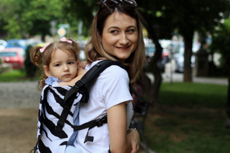 Marsupiu ergonomic Luna Una de culoare neagra, purtat de un părinte cu bebeluș. Design ajustabil, bretele moi și glugă pentru susținerea capului. [1]