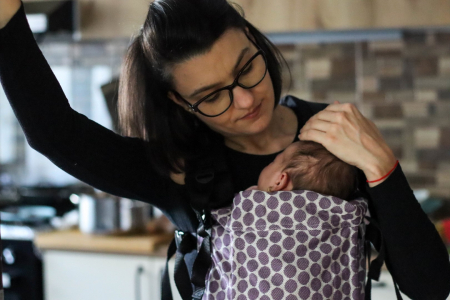 Marsupiu ergonomic Luna Una de culoare neagra, purtat de un părinte cu bebeluș. Design ajustabil, bretele moi și glugă pentru susținerea capului. [1]