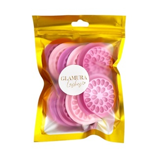 Suport din plastic roz în formă de floare cu multiple cavități circulare pentru adeziv și margini crestate, așezat pe o masă de lucru lângă o cutie de extensii de gene. [1]