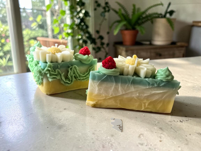 Lumânare artizanală din ceară de soia în formă de felie de tort, stratificată în galben, alb și turcoaz, decorată cu frișcă verde, o margaretă albă și o zmeură. [3]