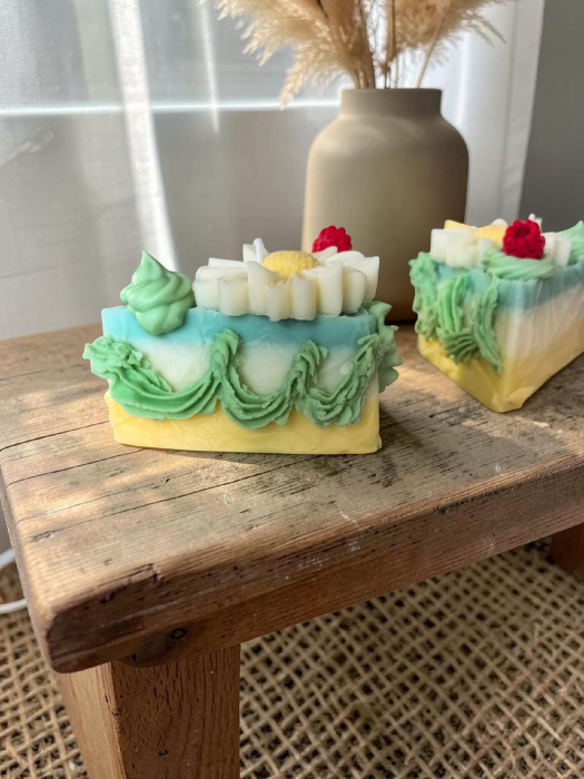 Lumânare artizanală din ceară de soia în formă de felie de tort, stratificată în galben, alb și turcoaz, decorată cu frișcă verde, o margaretă albă și o zmeură. [4]