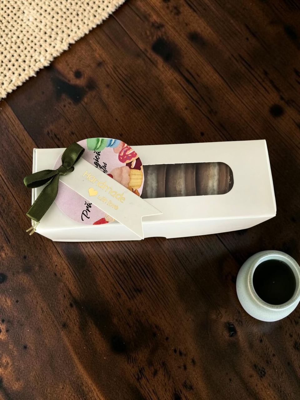 Cutie albă decupată care conține cinci pastile din ceară de soia parfumată modelate sub formă de prăjituri macarons maro cu cremă albă, așezate pe un pat de hârtie decorativă. [2]