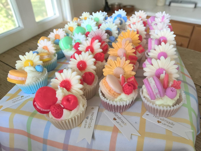 O colecție amplă de lumânări artizanale tip cupcake, în diverse culori vibrante, decorate cu flori margarete, fructe de pădure și macarons din ceară. [5]