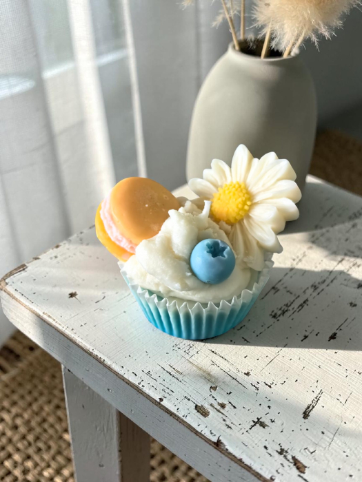 O colecție amplă de lumânări artizanale tip cupcake, în diverse culori vibrante, decorate cu flori margarete, fructe de pădure și macarons din ceară. [2]