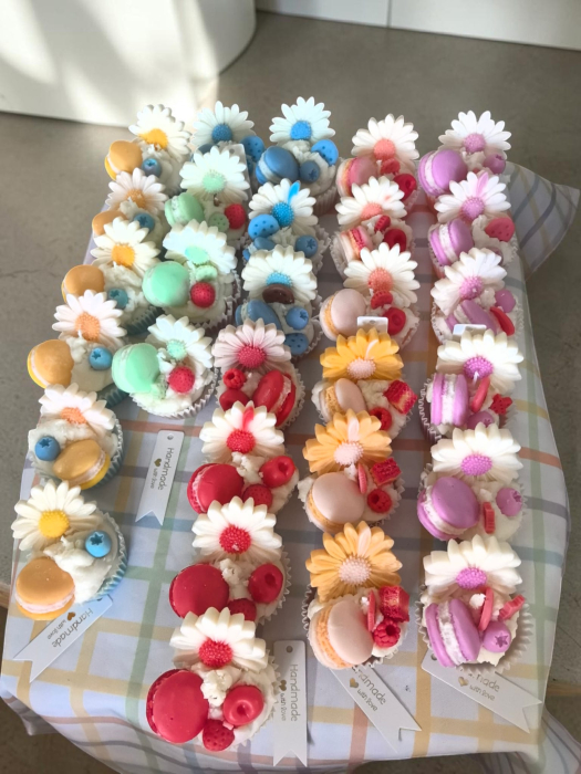 O colecție amplă de lumânări artizanale tip cupcake, în diverse culori vibrante, decorate cu flori margarete, fructe de pădure și macarons din ceară. [4]