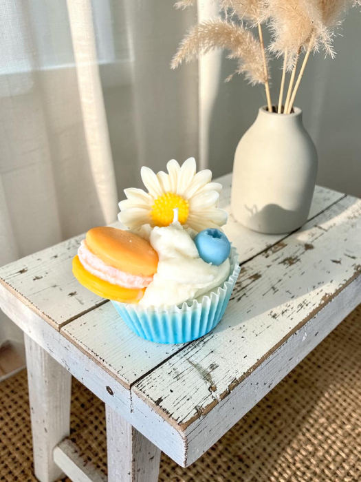 O colecție amplă de lumânări artizanale tip cupcake, în diverse culori vibrante, decorate cu flori margarete, fructe de pădure și macarons din ceară. [3]