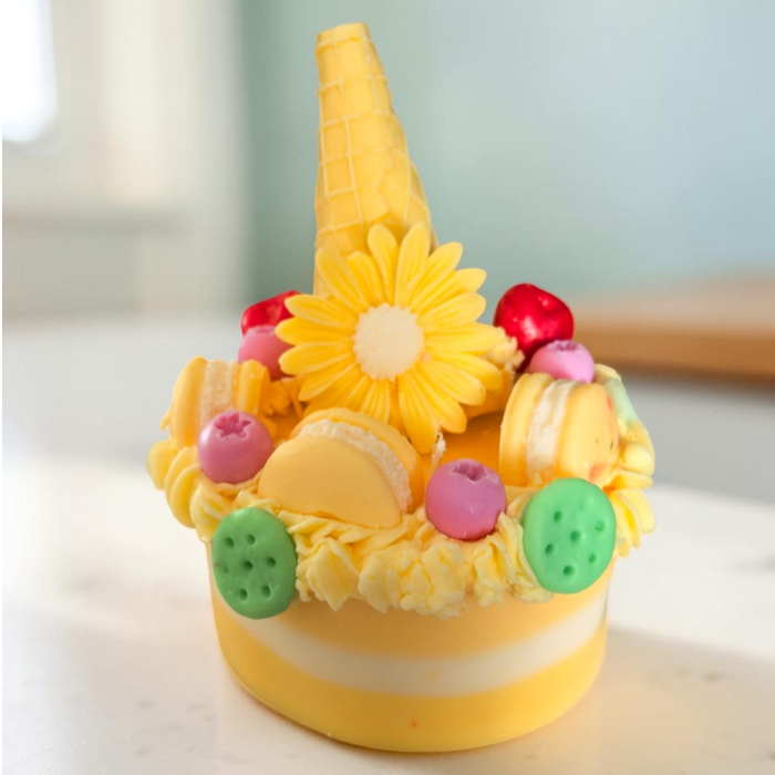 Lumânare din ceară de soia sub formă de tort cu dungi galbene și albe, decorată cu frișcă din ceară, cornet, floare, macarons și bile colorate. [3]