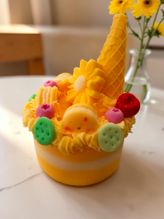 Lumânare din ceară de soia sub formă de tort cu dungi galbene și albe, decorată cu frișcă din ceară, cornet, floare, macarons și bile colorate. [2]