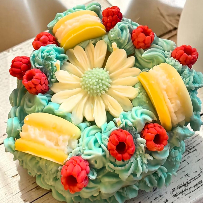 Lumânare din ceară de soia sub formă de tort, cu bază galbenă și glazură turcoaz, decorată cu margaretă, zmeură roșie și macarons galbene. [3]