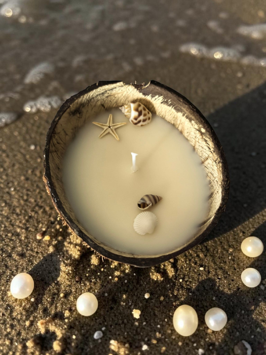 Lumânare parfumată din ceară de soia naturală turnată într-o nucă de cocos, decorată cu scoici și o steluță de mare, așezată pe nisipul plajei. [2]