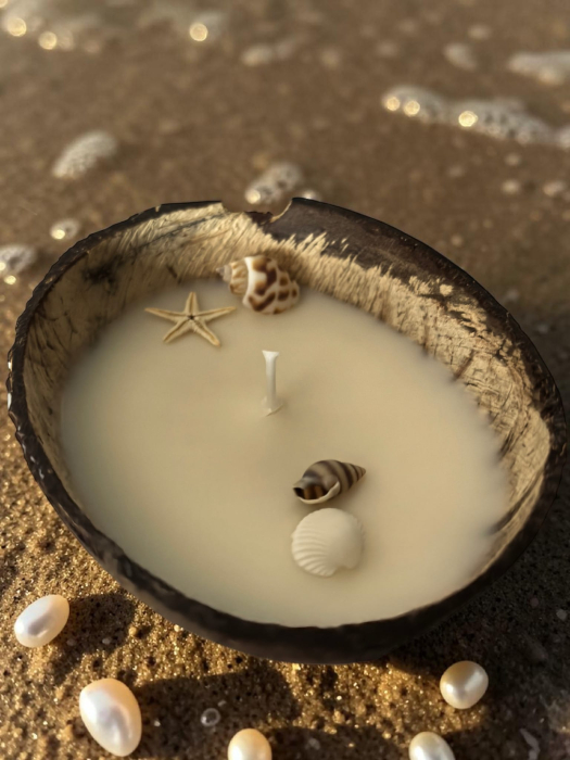 Lumânare parfumată din ceară de soia naturală turnată într-o nucă de cocos, decorată cu scoici și o steluță de mare, așezată pe nisipul plajei. [4]