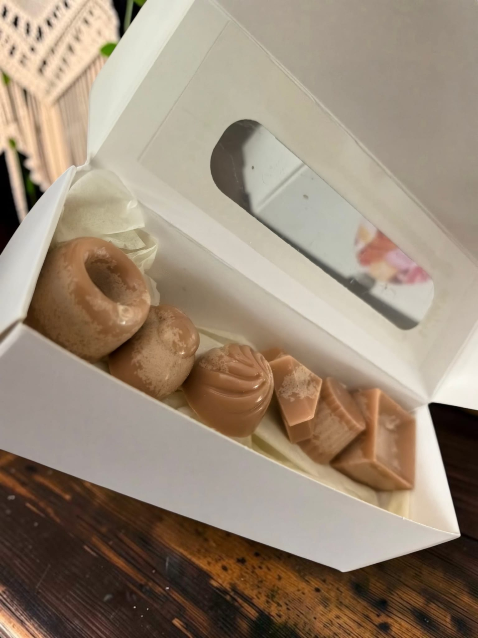 Cutie albă deschisă ce conține șase pastile de ceară parfumată (wax melts) din ceară de soia, cu design hiper-realist de praline de ciocolată în diferite forme geometrice. [2]