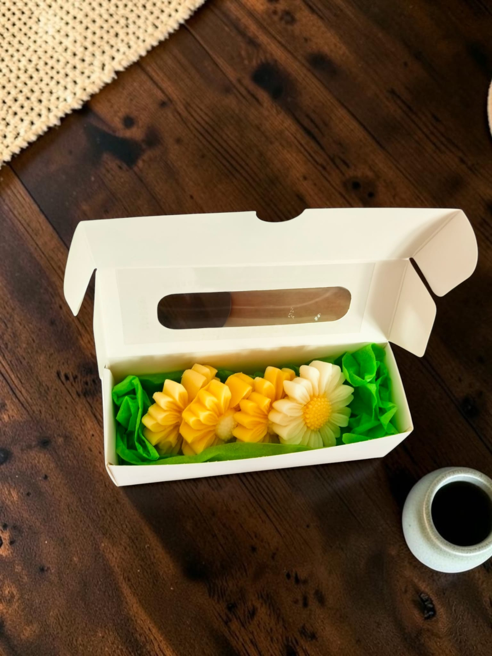 Cutie albă din carton cu fereastră transparentă, în care sunt așezate pastile de ceară parfumată în formă de margaretă. Cutia este decorată pe lateral cu o panglică verde și o etichetă rotundă cu mesajul "Handmade with love". [2]