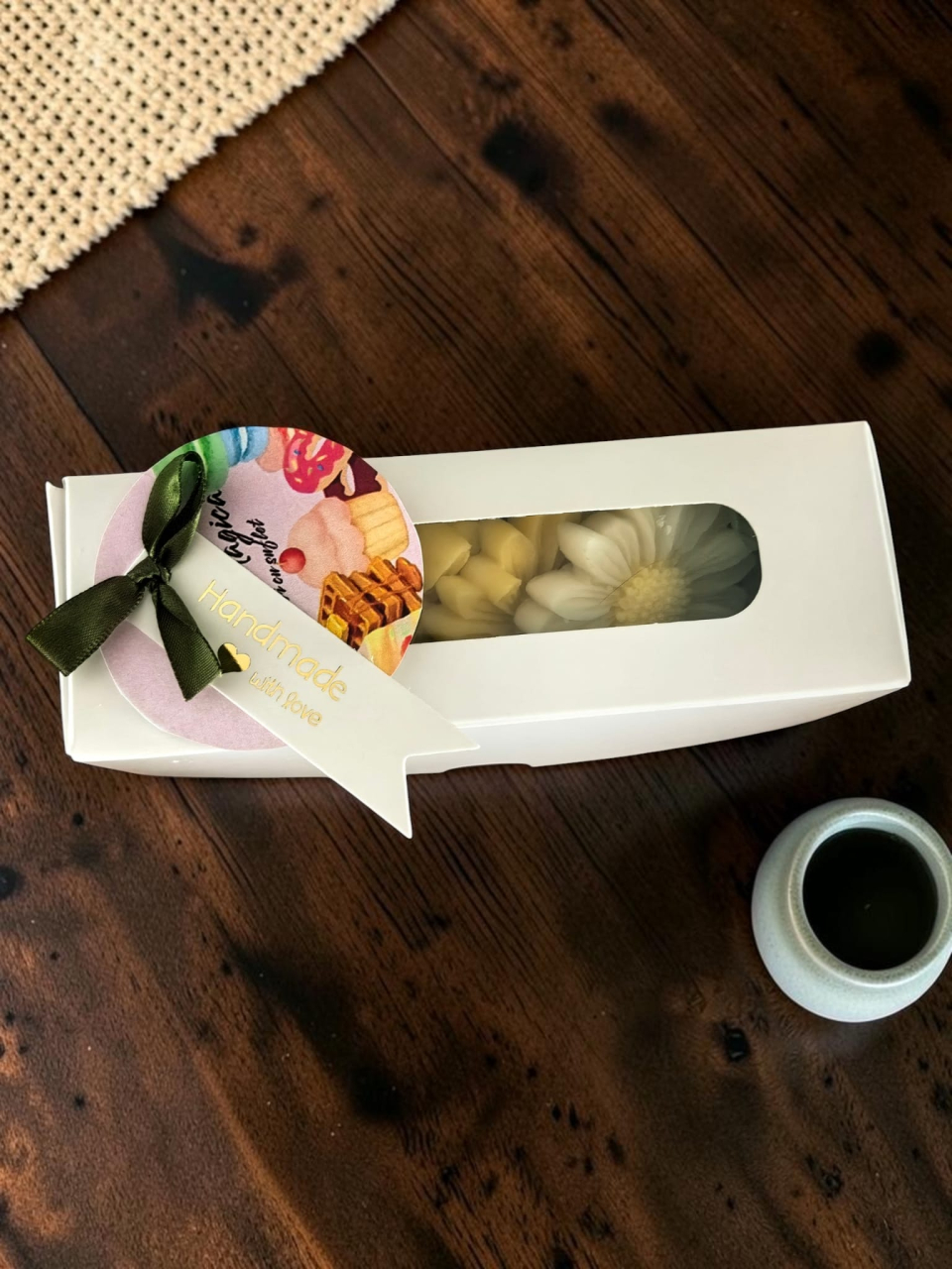 Cutie albă din carton cu fereastră transparentă, în care sunt așezate pastile de ceară parfumată în formă de margaretă. Cutia este decorată pe lateral cu o panglică verde și o etichetă rotundă cu mesajul "Handmade with love". [3]