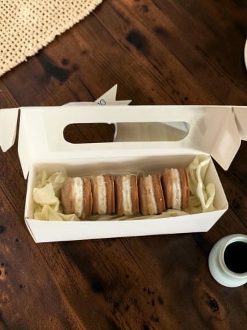 Delicii Parfumate: Lumânări Desert - Cutie albă decupată care conține cinci pastile din ceară de soia parfumată modelate sub formă de prăjituri macarons maro cu cremă albă, așezate pe un pat de hârtie decorativă.