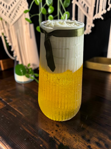 Delicii Parfumate: Lumânări Desert - Lumânare artizanală în pahar înalt de sticlă striată cu aspect de halbă de bere, bază galbenă din ceară gel și spumă albă din ceară de soia decorată cu panglică verde.
