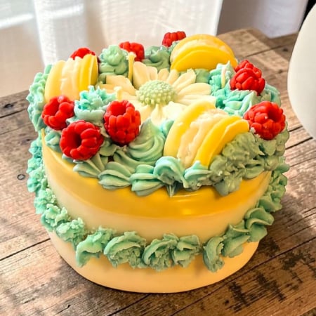 Delicii Parfumate: Lumânări Desert - Lumânare din ceară de soia sub formă de tort, cu bază galbenă și glazură turcoaz, decorată cu margaretă, zmeură roșie și macarons galbene.