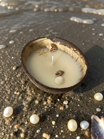 Lumânări parfumate lucrate manual - Lumânare parfumată din ceară de soia naturală turnată într-o nucă de cocos, decorată cu scoici și o steluță de mare, așezată pe nisipul plajei.