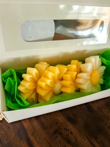 Delicii Parfumate: Lumânări Desert - Cutie albă din carton cu fereastră transparentă, în care sunt așezate pastile de ceară parfumată în formă de margaretă. Cutia este decorată pe lateral cu o panglică verde și o etichetă rotundă cu mesajul "Handmade with love".